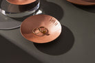 A small, round, matte copper trinket dish with an seven tally engraved along the rim. The dish has been photographed atop a moss green background. Within in the trinket dish there is two gold rings, one with a large amber stone. The trinket dish is made by Empire Copper.